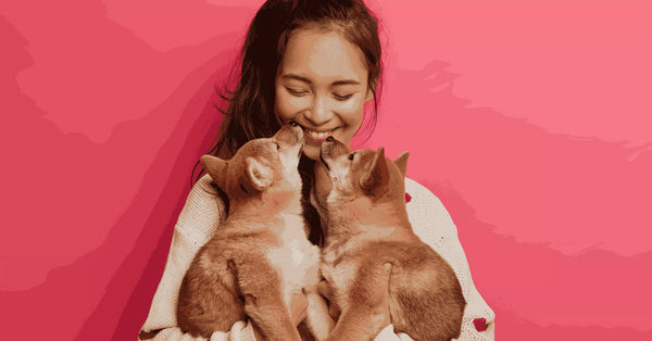Smiling woman cuddling two adorable puppies against a pink background, showing the loving bond between a pet mom and her dogs